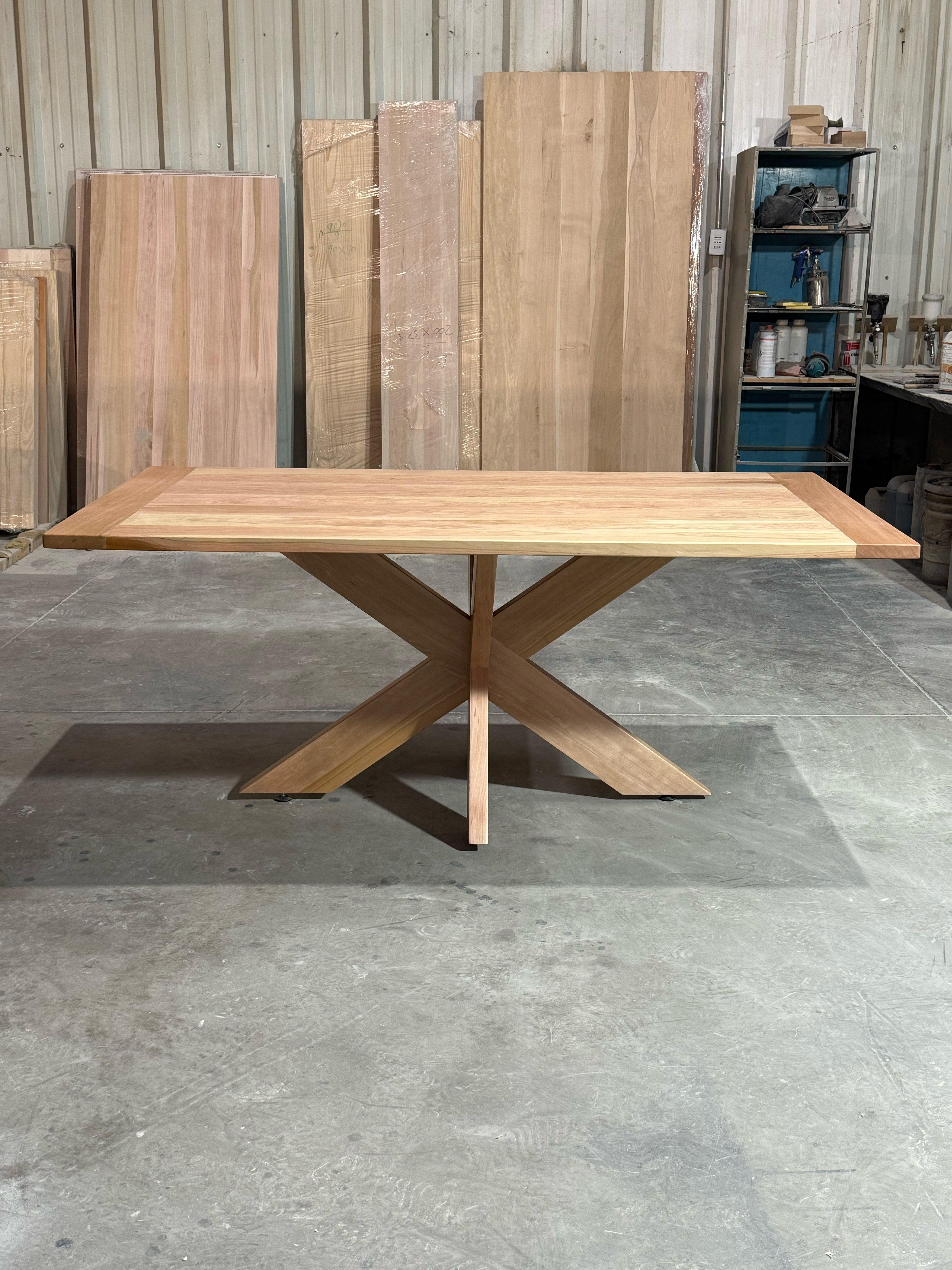 Vista frontal de la mesa de comedor rectangular Lena en lenga natural, con la pata principal de la base que forma una X a lo largo de la mesa.