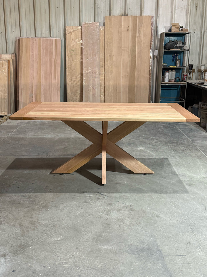 Vista frontal de la mesa de comedor rectangular Lena en lenga natural, con la pata principal de la base que forma una X a lo largo de la mesa.