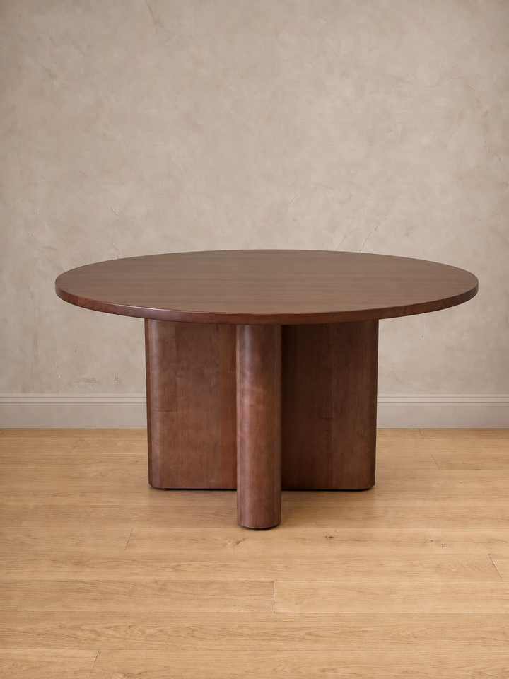 Mesa de comedor redonda Amora en tono nogal, con su base en cruz de remates curvos, en un comedor de muro claro y piso de madera.