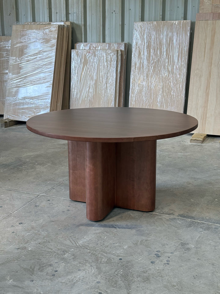 Mesa de comedor redonda Amora en lenga tono nogal, vista en tres cuartos con cubierta y base en cruz completas, en el taller de Santino Design.
